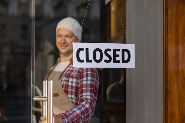 Beau propriétaire de bar debout près du panneau FERMÉ qui pend à la porte du café