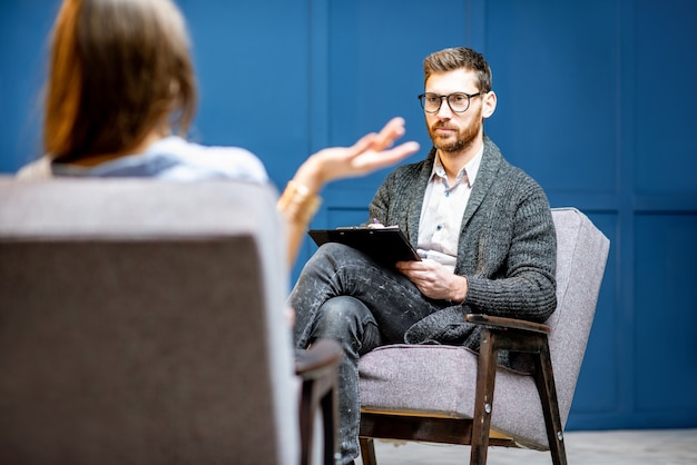 Beau psychologue masculin écoutant la cliente assise pendant une séance psychologique à l'intérieur du bureau bleu