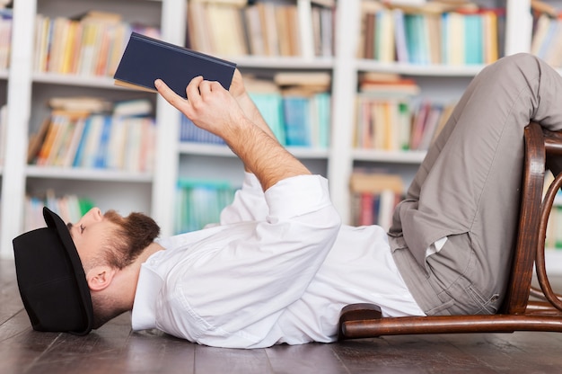 Beau rat de bibliothèque. Vue latérale d'un jeune homme réfléchi en chemise et bretelles allongé sur le sol et lisant un livre