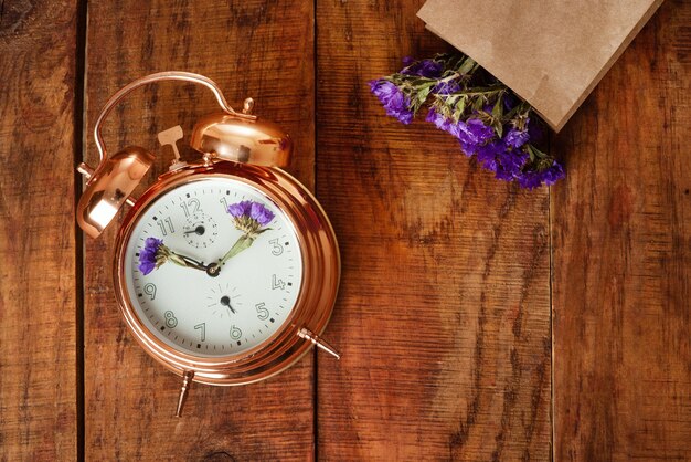 Beau réveil vintage en métal doré avec des fleurs bleues au lieu des aiguilles de l'horloge et un sac en papier avec des fleurs sur la surface en bois Awakening Spring time Temps pour le printemps Pic conceptuel