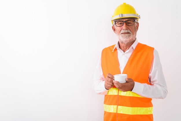 Beau senior homme barbu travailleur de la construction tenant une tasse de café tout en portant des lunettes sur blanc