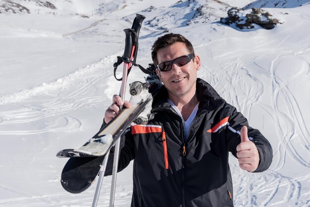 Beau skieur avec ses skis sur son épaule en regardant la caméra et en levant les pouces