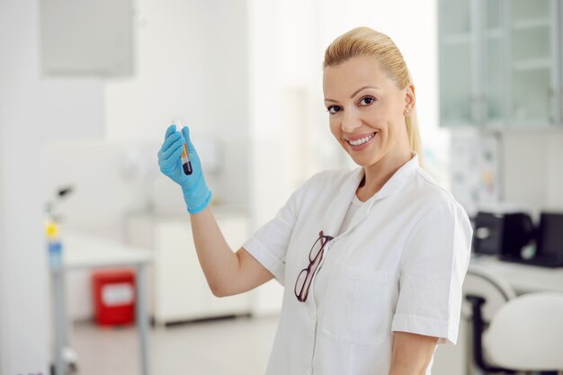 Beau sourire blond assistant de laboratoire debout en laboratoire et tenant un tube à essai avec du sang