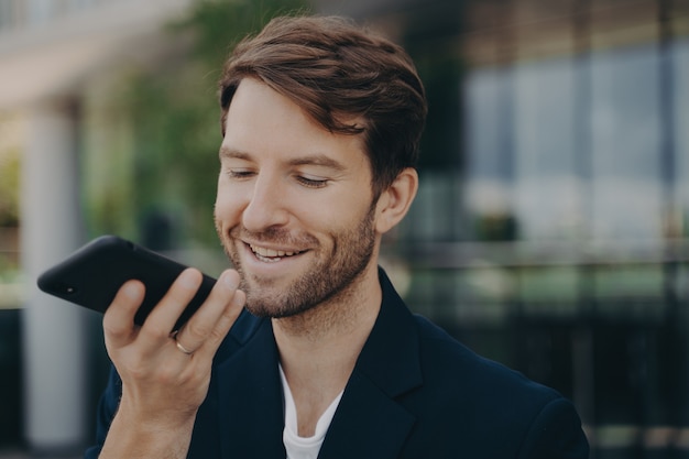 Beau sourire soigné employé de bureau barbu parlant sur haut-parleur debout à l'extérieur