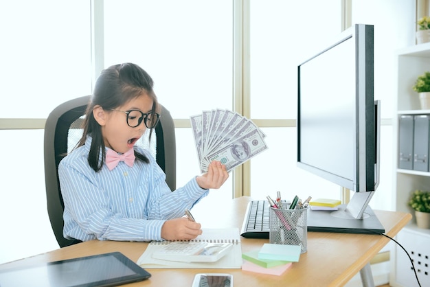 beau travail de bureau de petite fille douce tenant de nombreux billets de banque et utilisant un enregistrement de stylo sur le livre lorsqu'elle travaille au bureau de l'entreprise.