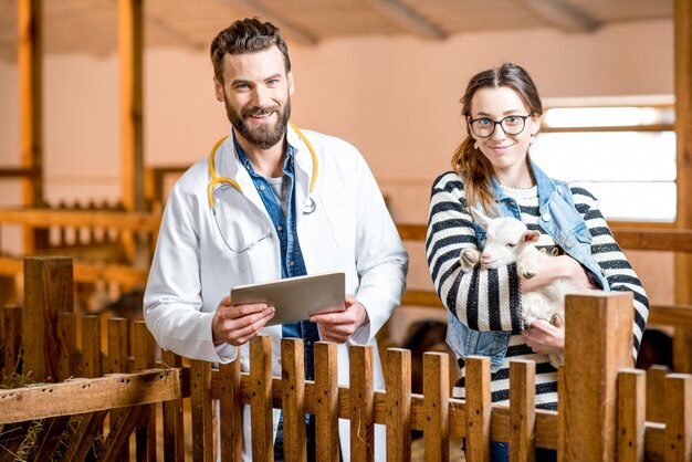 Beau vétérinaire en blouse médicale et jeune femme prenant soin du bébé chèvre debout à l'intérieur de la grange