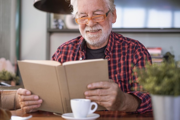 Un beau vieil homme barbu assis à une table de café en train de lire un livre en profitant d'une tasse de café espresso