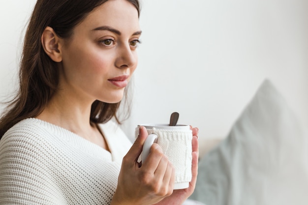 Beau visage d'une fille, une fille en pull blanc est assise sur un canapé avec une tasse de thé chaud dans les mains