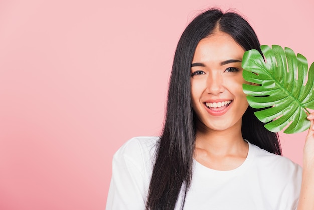 Beau visage. Portrait d'une belle jeune femme asiatique à la peau fraîche et saine tenant une feuille de monstère verte sur son visage, Feuilles tropicales, prise de vue en studio isolée sur fond rose, concept de spa pour les soins du corps de la peau