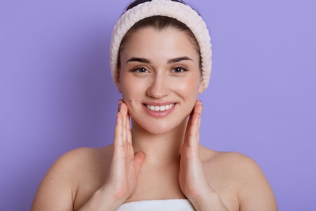 Beau visage portrait de jeune femme touchant ses joues et souriant tout en regardant directement, debout avec les épaules nues et porte un bandeau de cheveux. Soins de la peau et santé.