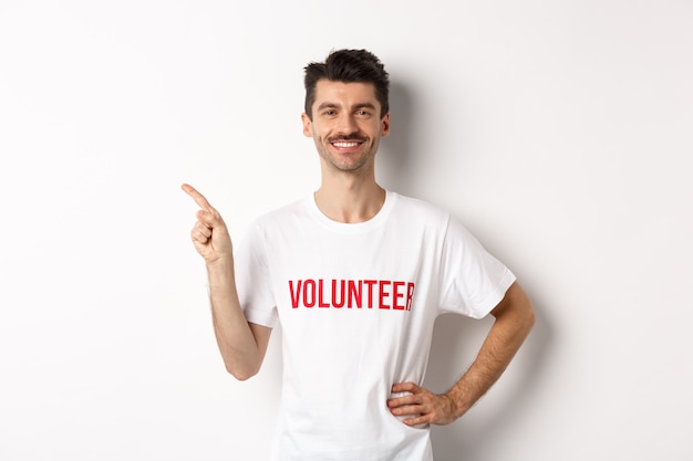 Beau volontaire souriant en t-shirt pointant le doigt vers la gauche, montrant l'annonce, debout sur fond blanc.