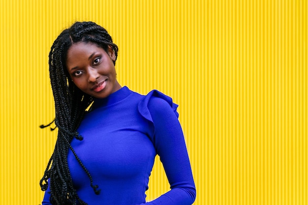 La beauté afro-américaine avec une coiffure afro tressée pose dans un mur jaune portant une tenue bleue colorée