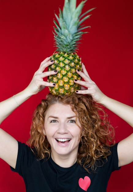 Beauté Aux Cheveux Roux Dans Un T-shirt Noir Avec Une Impression De Coeur Tenir Un Ananas Dans Ses Mains Sur Un Espace Rouge