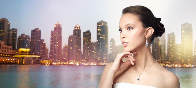 beauté, bijoux, accessoires de mariage, personnes et concept de luxe - belle femme asiatique ou mariée avec boucle d'oreille et pendentif sur fond de gratte-ciel et lumières de la ville de dubaï en soirée