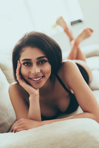 Beauté coquette. Belle jeune femme métisse en lingerie noire regardant la caméra et souriant en position couchée sur le canapé à la maison