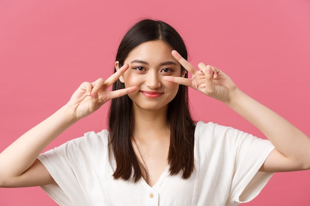 Beauté, émotions des gens et concept de loisirs d'été. Jolie fille japonaise mignonne et timide montrant des signes de paix kawaii près des yeux et souriant idiot à la caméra, debout sur fond rose.