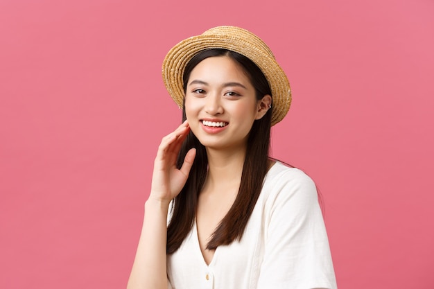 Beauté, émotions des gens et concept de loisirs et de vacances d'été. Joyeuse touriste asiatique profitant du week-end, regardant la caméra avec un large sourire joyeux, portant un chapeau de paille, fond rose.