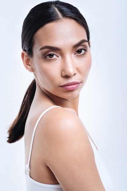 Beauté exotique. Le portrait d'une belle jeune femme aux cheveux noirs dans un débardeur blanc tout en se tenant à moitié tourné contre un mur blanc