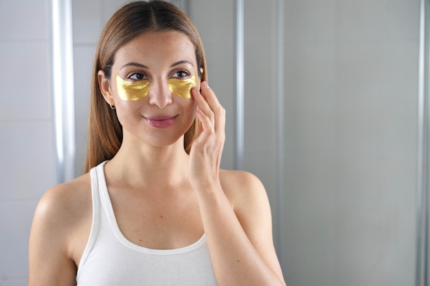 Photo beauté femme appliquant un masque anti-âge doré sous les yeux se regardant dans le miroir de la salle de bain soins de la peau fille toucher des patchs de masque en tissu sous les yeux pour réduire les poches sous les yeux