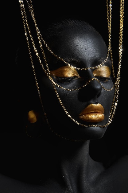 Beauté femme peinte en couleur de peau noire art corporel chaîne en or dans ses mains et autour de son cou Maquillage doré lèvres paupières bout des doigts ongles en peinture couleur or Maquillage professionnel