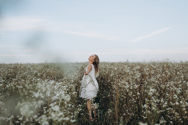 Beauté fille blonde aux yeux bleus et cheveux longs sur la tête, marchant dans le champ avec des fleurs blanches