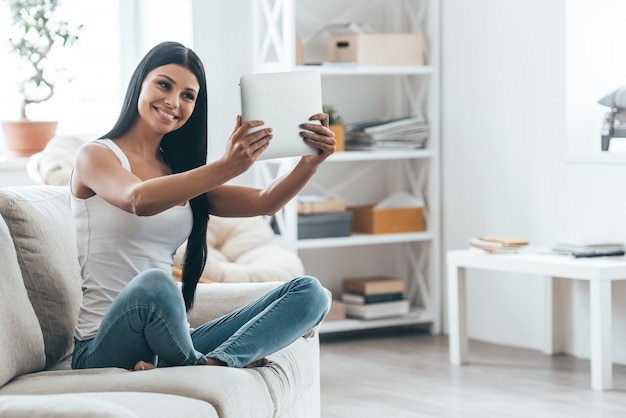 Beauté à la maison. Jolie jeune femme tenant une tablette numérique et souriante assise sur le canapé à la maison