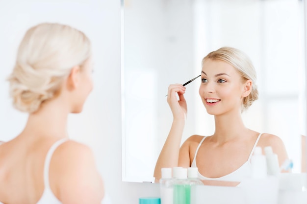 beauté, maquillage, cosmétiques, matin et concept de personnes - jeune femme se maquillant en dessinant des sourcils avec une brosse et regardant au miroir dans la salle de bain de la maison