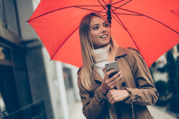 Beauté avec parapluie. Jolie jeune femme souriante portant un parapluie et un téléphone portable en se tenant debout dans la rue