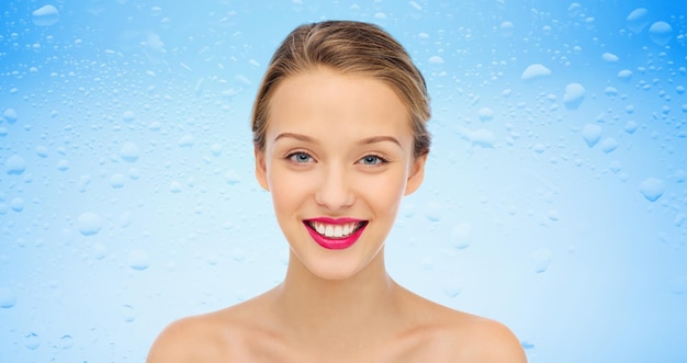 beauté, personnes, hydratation, soins de la peau et concept de santé - visage souriant de jeune femme avec rouge à lèvres rose sur les lèvres et les épaules au-dessus des gouttes d'eau sur fond bleu