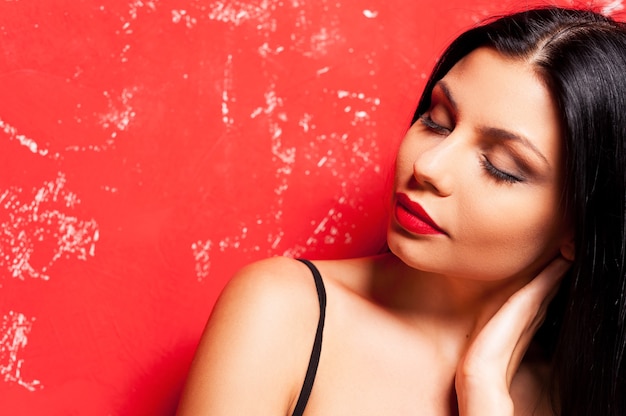 Beauté sur rouge. Portrait d'une belle jeune femme gardant les yeux fermés et touchant son cou avec la main en se tenant debout sur fond rouge