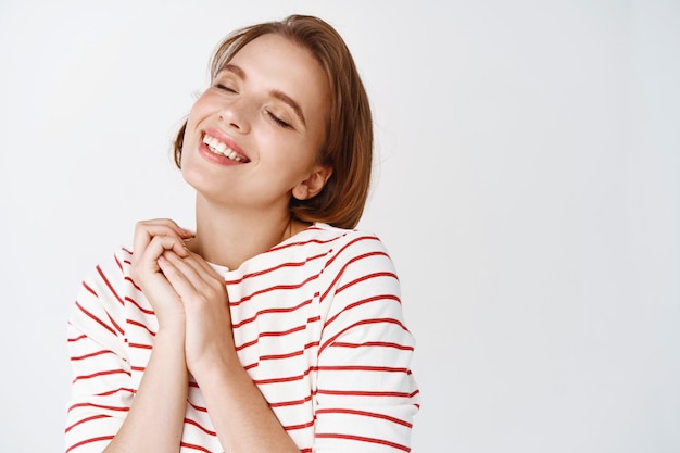 Beauté et soins de la peau. Heureuse jeune femme avec un maquillage à la lumière naturelle, cheveux courts, rêvant de quelque chose de doux, sourire tendrement et fermer les yeux, mur blanc