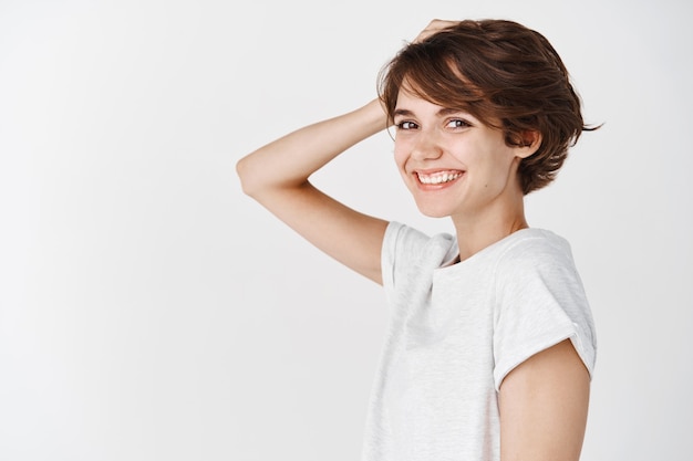 Beauté et soins de la peau. Portrait d'une jeune fille authentique avec une coiffure courte en désordre, des cheveux touchants et souriante heureuse debout sur un mur blanc