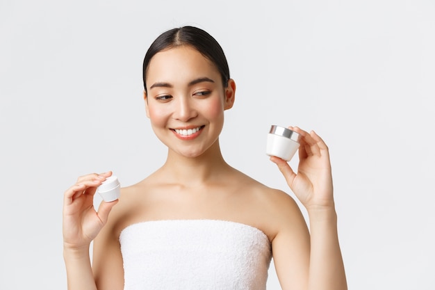 Beauté, soins personnels, salon spa et concept de soins de la peau. Gros plan de la belle femme asiatique en serviette de bain tenant deux crèmes, produits nourrissants pour les yeux et le visage, souriant, traitement de la peau