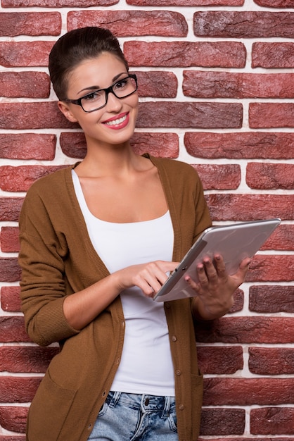 Beauté avec tablette numérique. Jolie jeune femme aux cheveux courts dans des verres travaillant sur une tablette numérique et souriante en se tenant debout contre le mur de briques