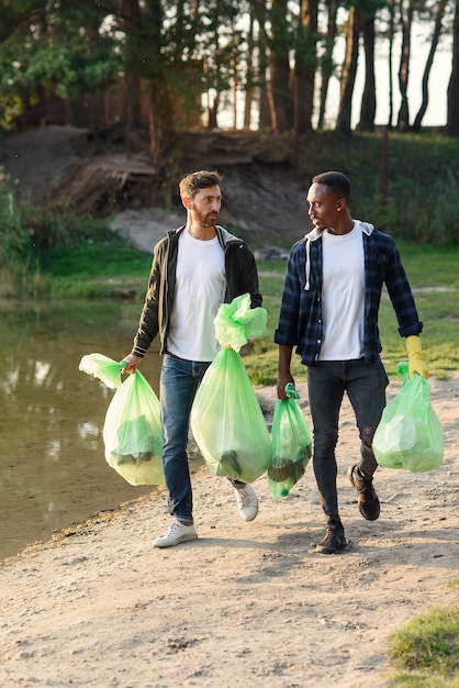 Beaux amis actifs métis transportant des sacs en plastique près du lac après le nettoyage du territoire environnant des ordures.