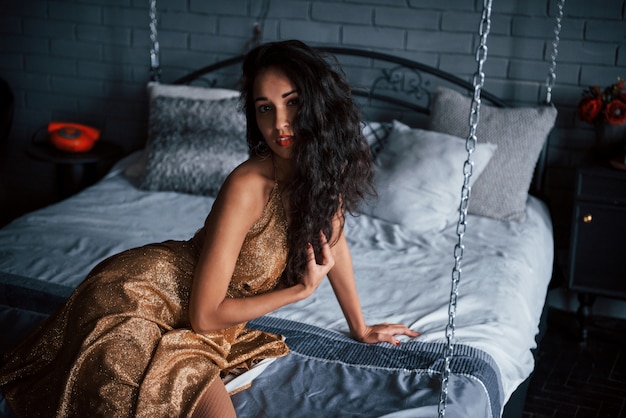 De beaux cheveux noirs. Fille en robe dorée est assise au lit blanc sur les chaînes dans les appartements de luxe