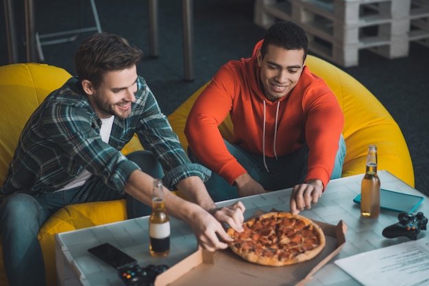 Beaux hommes affamés assis ensemble sur le canapé tout en prenant des morceaux de pizza