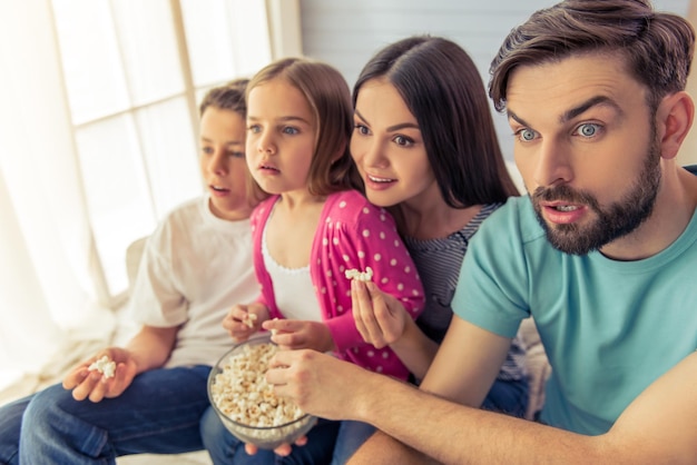 De beaux jeunes parents, leur fille et leur fils regardent la télévision, mangent du pop-corn et montrent la surprise, assis sur un canapé à la maison