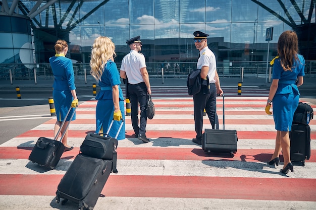 De beaux pilotes et de charmantes hôtesses de l'air portant des sacs à bagages à roulettes en marchant sur un passage pour piétons