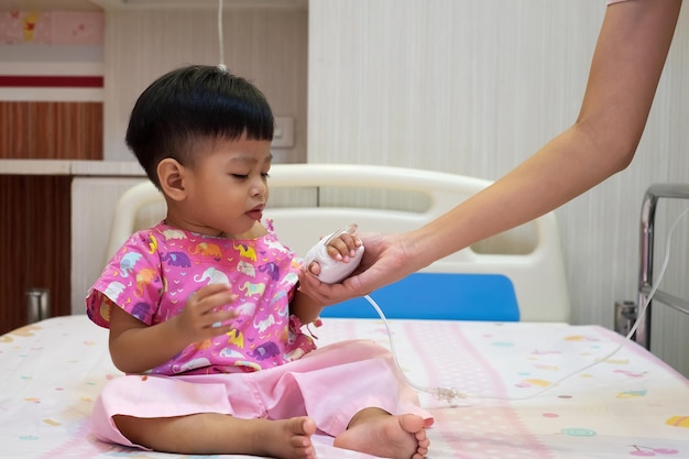 Un bébé de 2 ans vêtu de rose est assis sur un lit, tenant la main de sa mère à l'hôpital