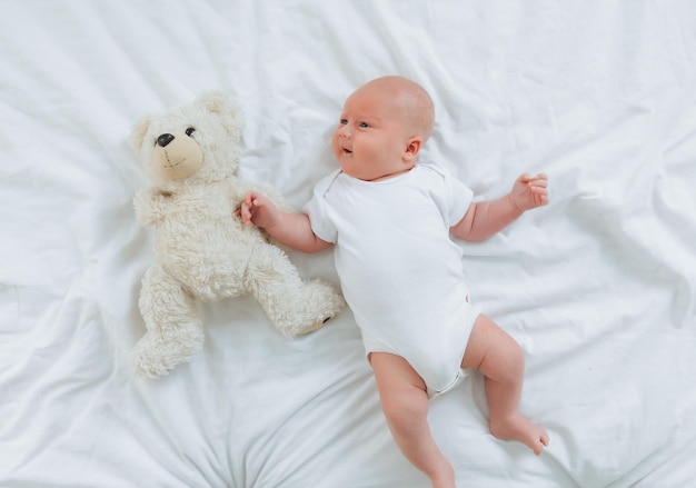 Un bébé de 2 mois dans un body blanc est allongé à la maison sur un lit blanc à côté d'un ours en peluche vue d'en haut nouveau-né