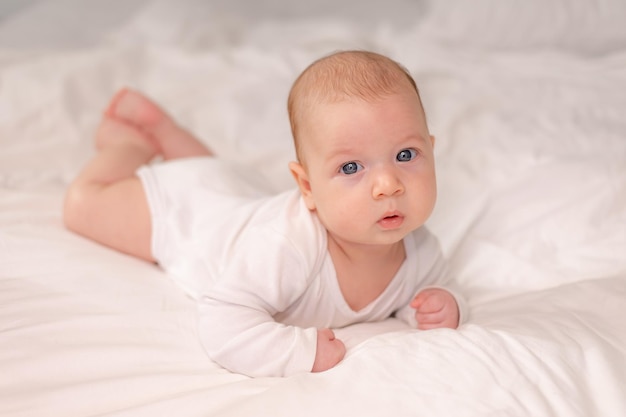 Bébé aux yeux bleus dans un body blanc allongé sur le ventre sur du linge de lit blanc