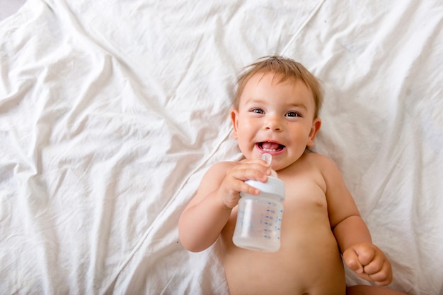 Bébé en bas âge allongé sur le lit blanc, sourit et boit de l&#39;eau d&#39;une bouteille en plastique