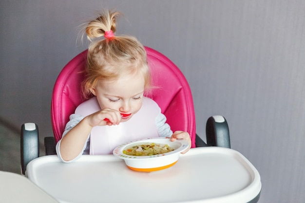 Bébé en bas âge avec un visage sale mangeant de la soupe elle-même avec une cuillère Jolie petite fille assise dans une chaise haute pour bébé dans la cuisine à la maison