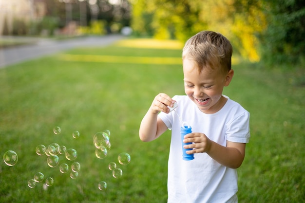 Le Bébé Blond Mignon Gonfle Des Bulles De Savon En été Sur Une Pelouse Verte, S'amusant, Loisirs De Plein Air