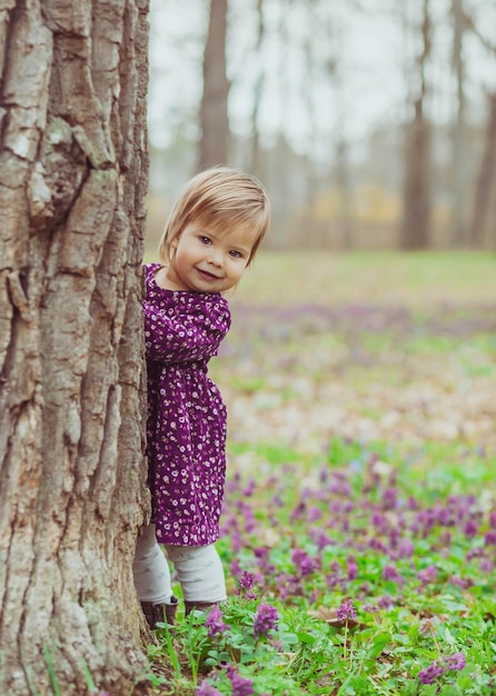 bébé blond vêtu d'une robe colorée sort de derrière un arbre dans la forêt