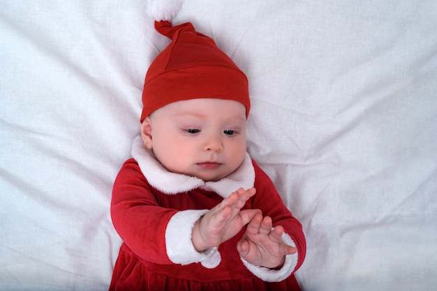 Bébé en bonnet de noel allongé sur un fond blanc, joue avec ses mains. Concept de Noël.