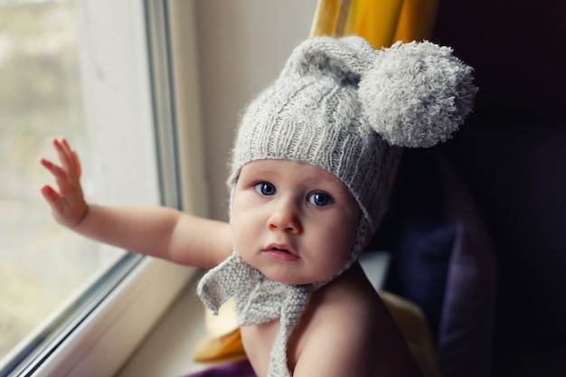 Un Bébé En Bonnet Tricoté S'assoit Sur Le Rebord De La Fenêtre Et Touche Le Verre, Veut Marcher, Danger