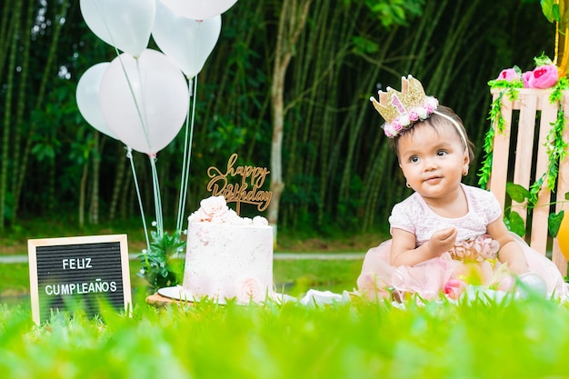 Bébé brunette latina assis sur la pelouse à l'air heureux de célébrer son premier anniversaire à côté d'un gâteau avec un signe de joyeux anniversaire