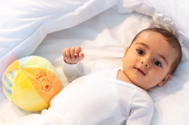 Bébé caucasien de 5 mois souriant dans un lit blanc vient de se réveiller, un body blanc et une balle pour jouer